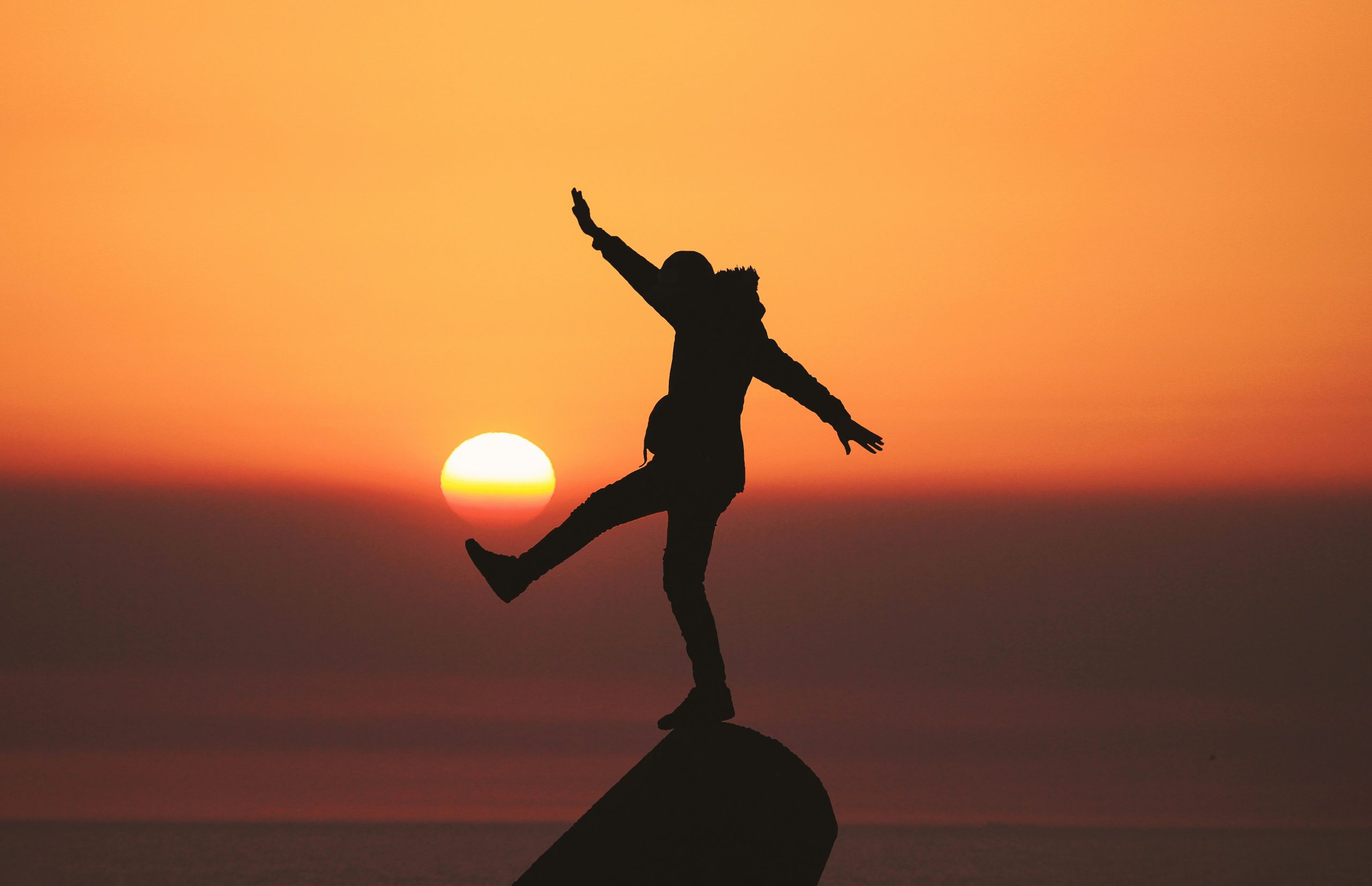 Silhouette of a person balancing on one leg on a rocky outcrop, arms outstretched, against an orange sunset over the sea